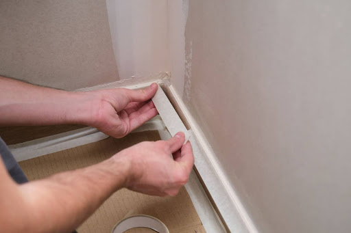 Painter applying masking tape along baseboards to prepare walls for a clean and precise paint job